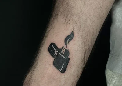 Tatouage blackwork d'un briquet Zippo stylisé et allumé, situé sur l'avant-bras d'une personne.