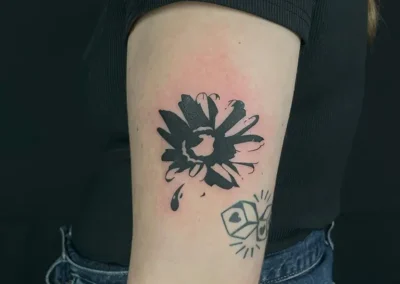 Tatouage blackwork d'une fleur épanouie avec des détails de style éclaboussure et une goutte, situé sur le bras d'une personne portant un t-shirt noir et un jean.