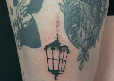 Tatouage blackwork d'une lanterne suspendue sur la cuisse, entourée d'autres tatouages dans un style similaire, dont un visage de femme et un motif ornemental.