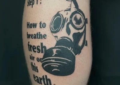 Tatouage blackwork sur un mollet, représentant un masque à gaz vintage détaillé à côté du texte "Step 1: How to breathe fresh air on this earth
