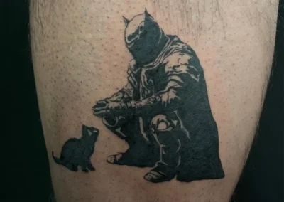Tatouage de Batman accroupi avec un petit chat noir à ses côtés, style bande dessinée ou illustratif en noir et gris, sur la jambe