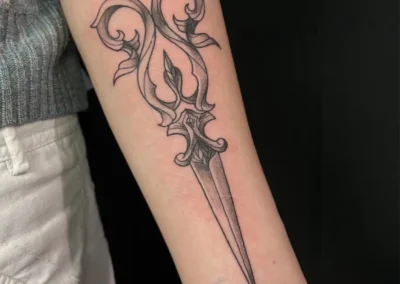 Tatouage d'une dague ornementale avec des volutes et des motifs floraux, style blackwork ou gothique, sur l'avant-bras