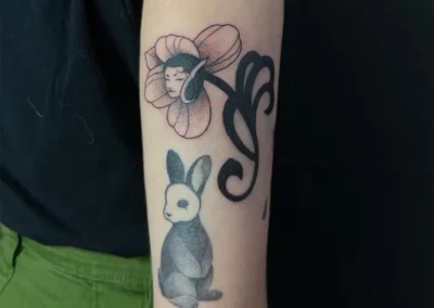 Tatouage d'une fleur avec un visage de femme endormi en son centre, et un lapin en dessous, style illustration ou cartoon en noir et gris, sur le bras