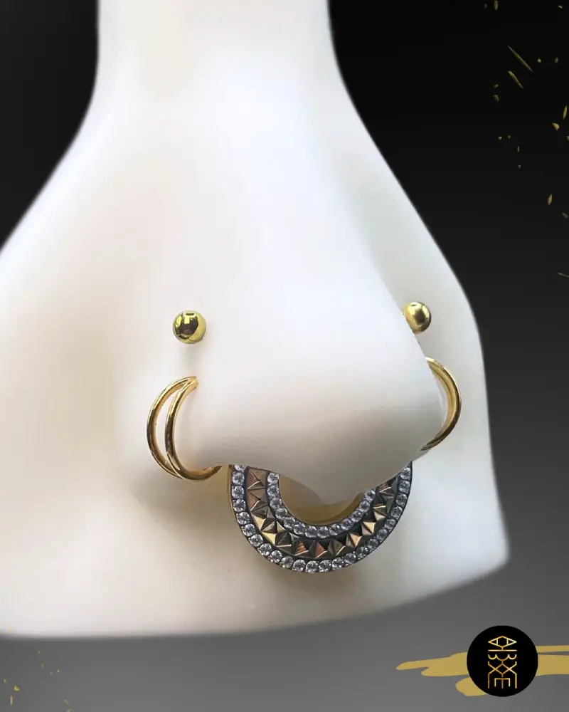 Bijoux de pose piercing Un présentoir de piercing avec un nez factice blanc portant des bijoux dorés : deux piercings de narine anneaux et un piercing septum en demi-cercle avec des strass.
