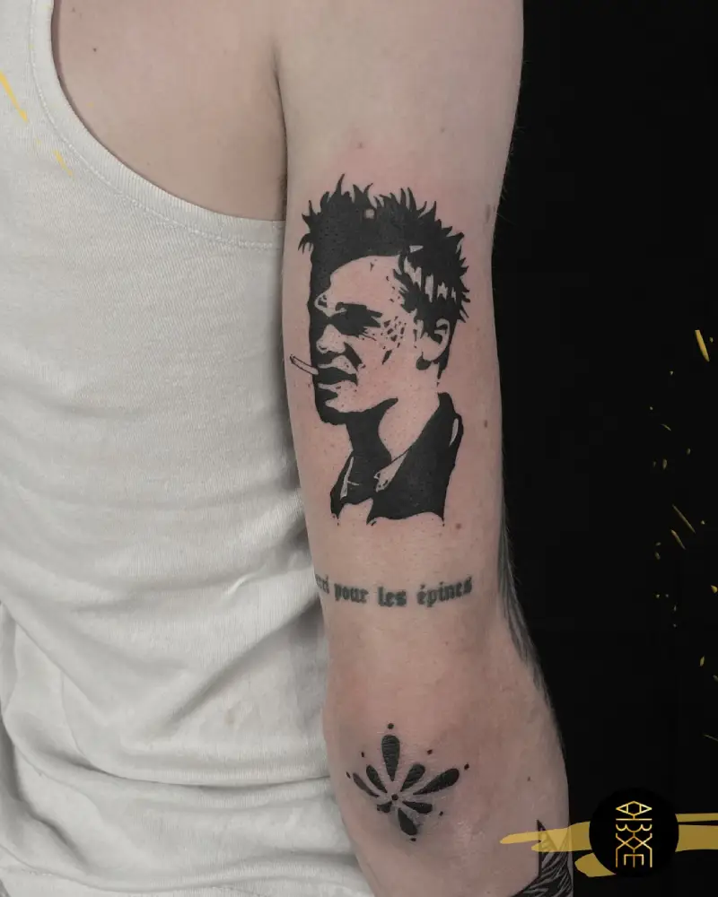 Cicatrisation tatouage Gros plan d'un bras avec un tatouage de visage masculin en noir et blanc, style réaliste ou portrait, avec une cigarette à la bouche, des cheveux ébouriffés et un regard intense. Sous le tatouage, on peut voir une partie de texte en français 'pour les épines' et un motif abstrait au niveau du coude.