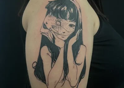 Tatouage d'une femme avec un double visage ou un visage déformé, avec une expression à la fois enfantine et inquiétante, dans un style manga en noir et gris sur l'épaule