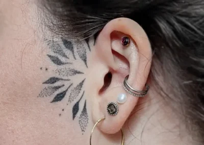 Tatouage ornemental et géométrique noir sur la joue, près de l'oreille. Lignes symétriques et dotwork